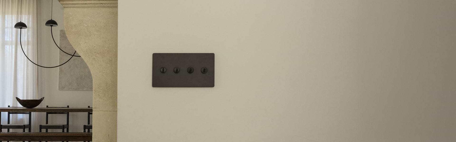 A minimalist light switch panel with four black switches on a cream wall, adjacent to a rustic dining area with a chandelier.