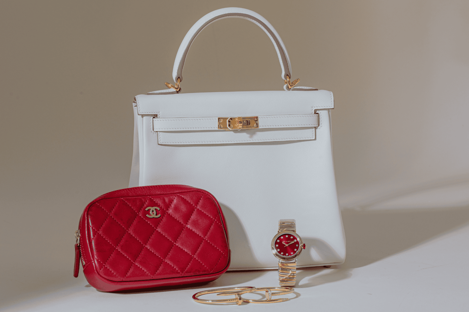 White handbag, red quilted pouch, gold watch, and gold chain on a beige background