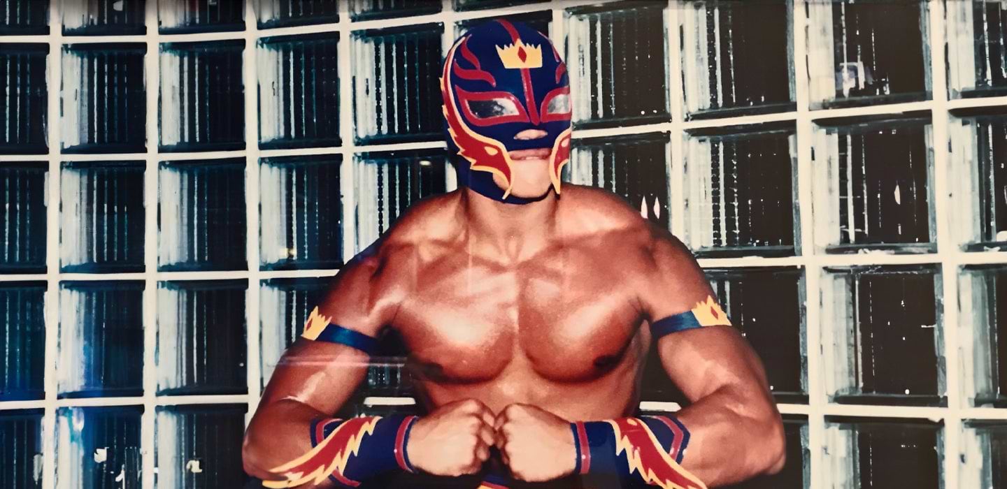 A masked wrestler in colorful gear poses in front of a glass block wall.