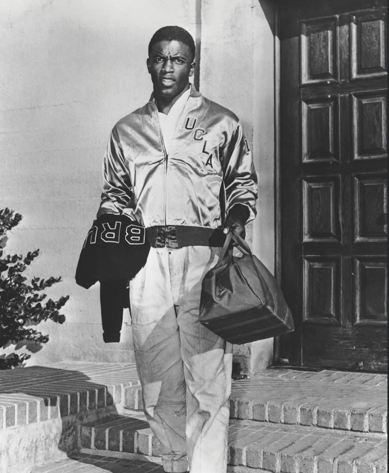 Man in a UCLA jacket holding a gym bag, standing outside a building.