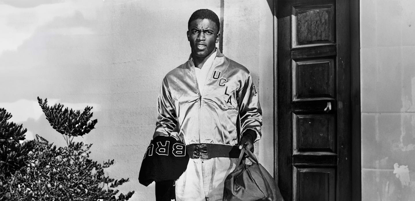 A man in a 'UCLA' jacket stands next to a door holding a duffle bag.