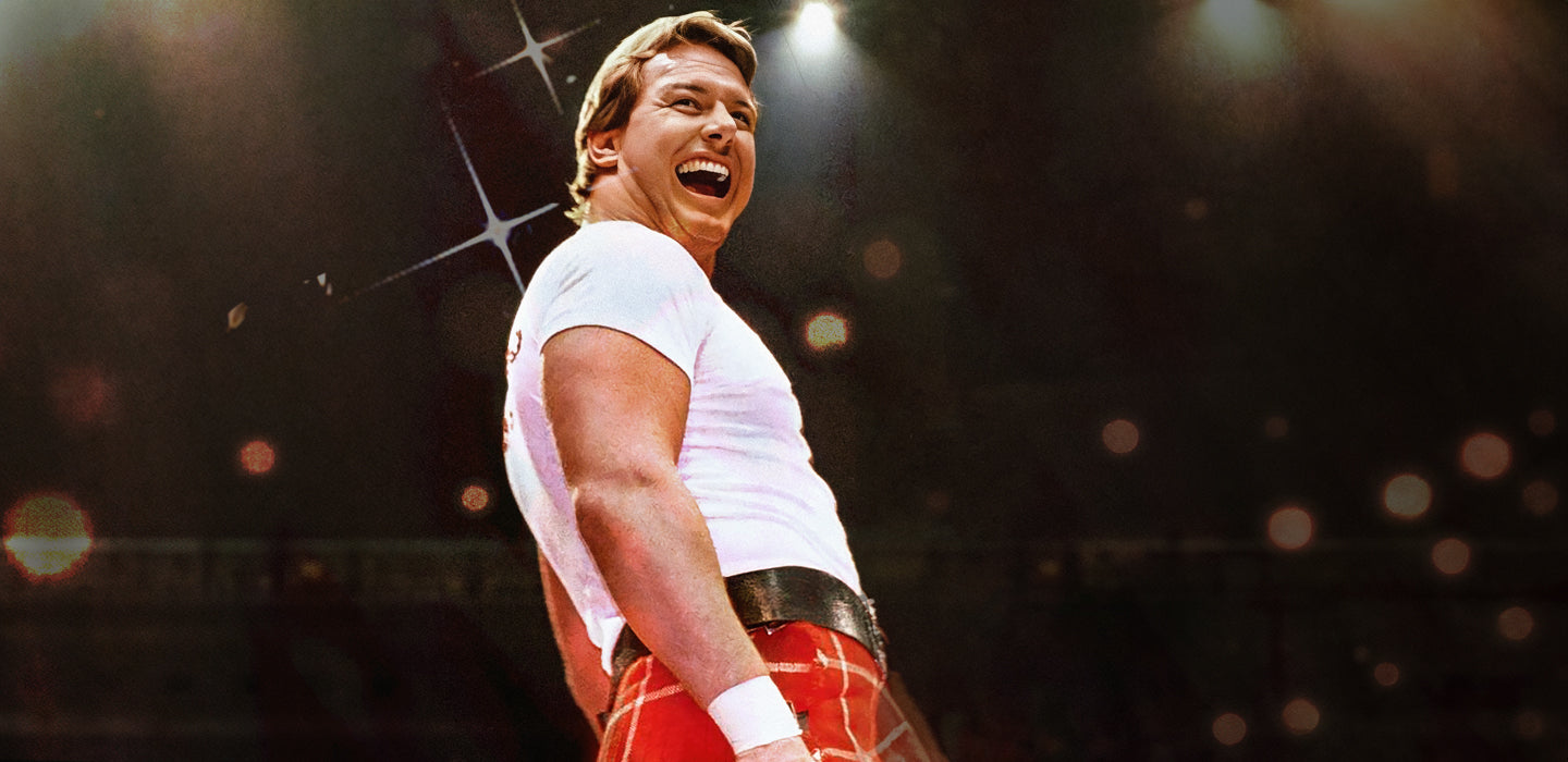 Man in a white shirt and red plaid kilt, smiling under spotlights.