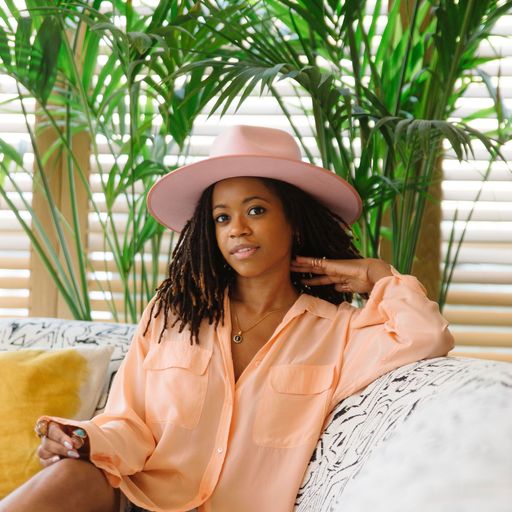 Chelsea VonChaz sits on a sofa in front of large house plants in a bright, sunny room