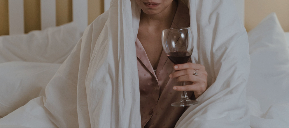 Person wrapped in a white blanket holding a glass of red wine while sitting on a bed.