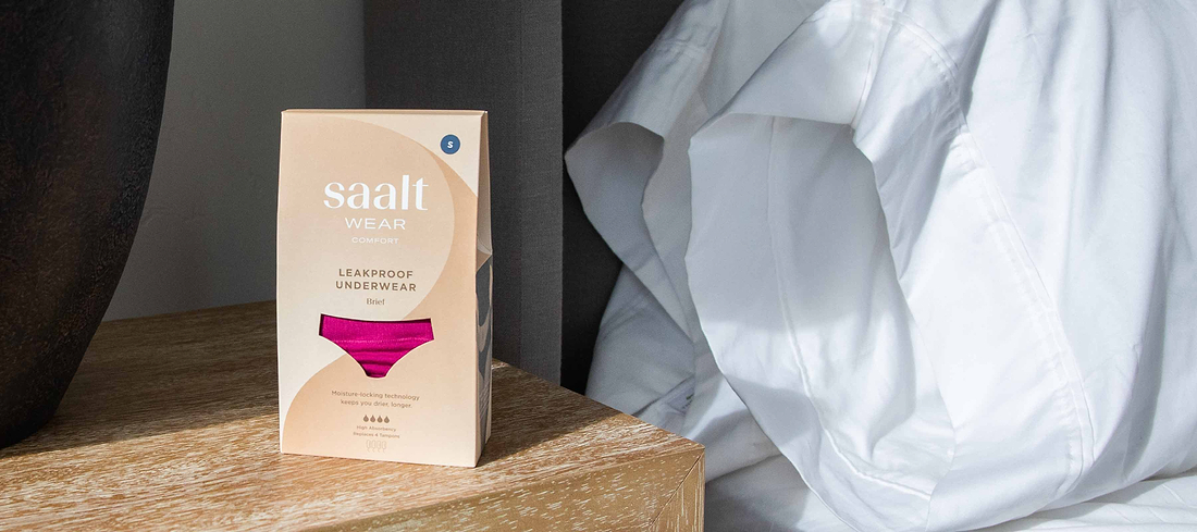 Saalt Wear leakproof underwear packaging on a bedside table beside white bedding and curtains.