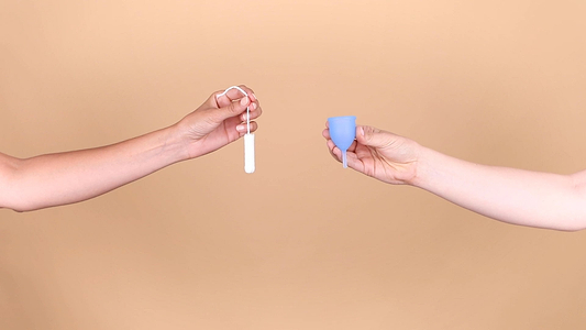 Hands holding a tampon and reusable menstrual cup against a beige background.