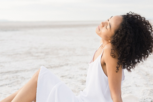 Woman in a white dress relaxing outdoors with eyes closed and curly hair blowing gently.