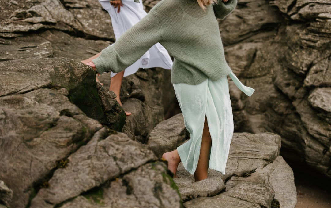 Person in oversized sweater and flowing skirt walking barefoot across rocky shoreline.