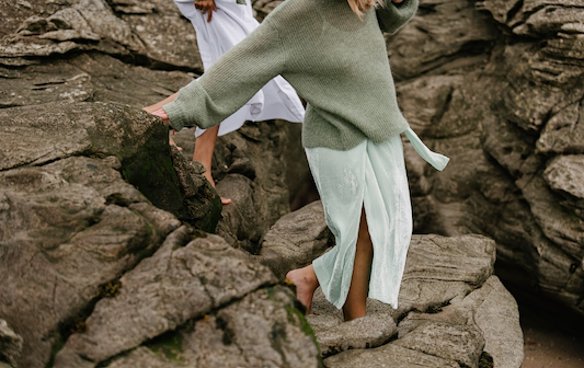 Two women carefully walking barefoot across rocky coastal terrain outdoors