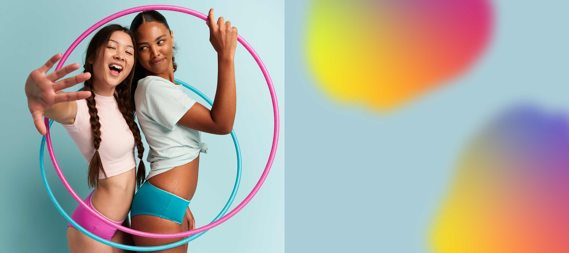 Two models wearing Saalt Teen period underwear in the style Brief. One is wearing pink, one blue. They are standing inside of two hula hoops, one pink one blue.