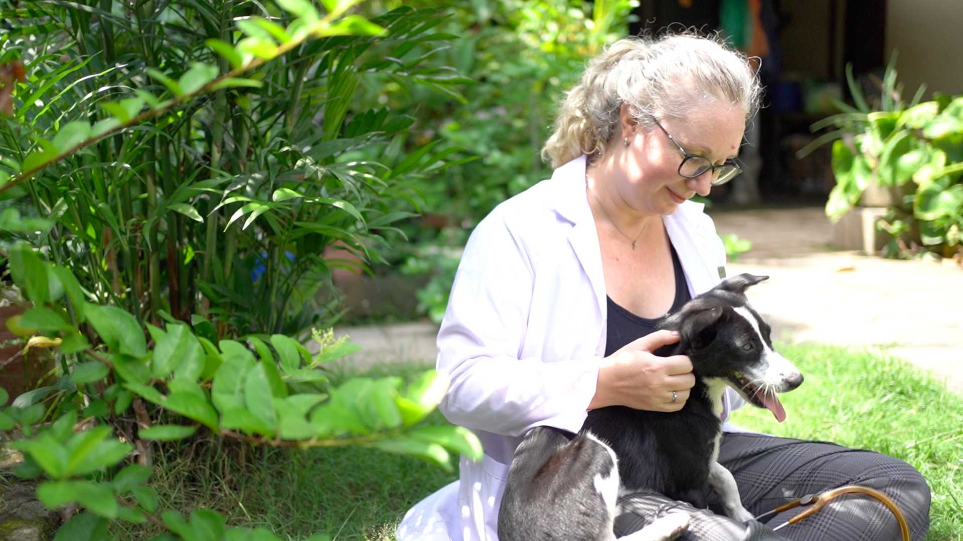 A woman sitting on the grass, smiling and petting a black and white dog.
