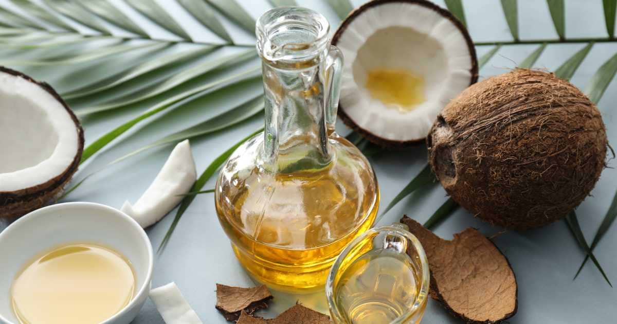 Coconut oil in a glass jug with whole and halved coconuts on a palm leaf background.