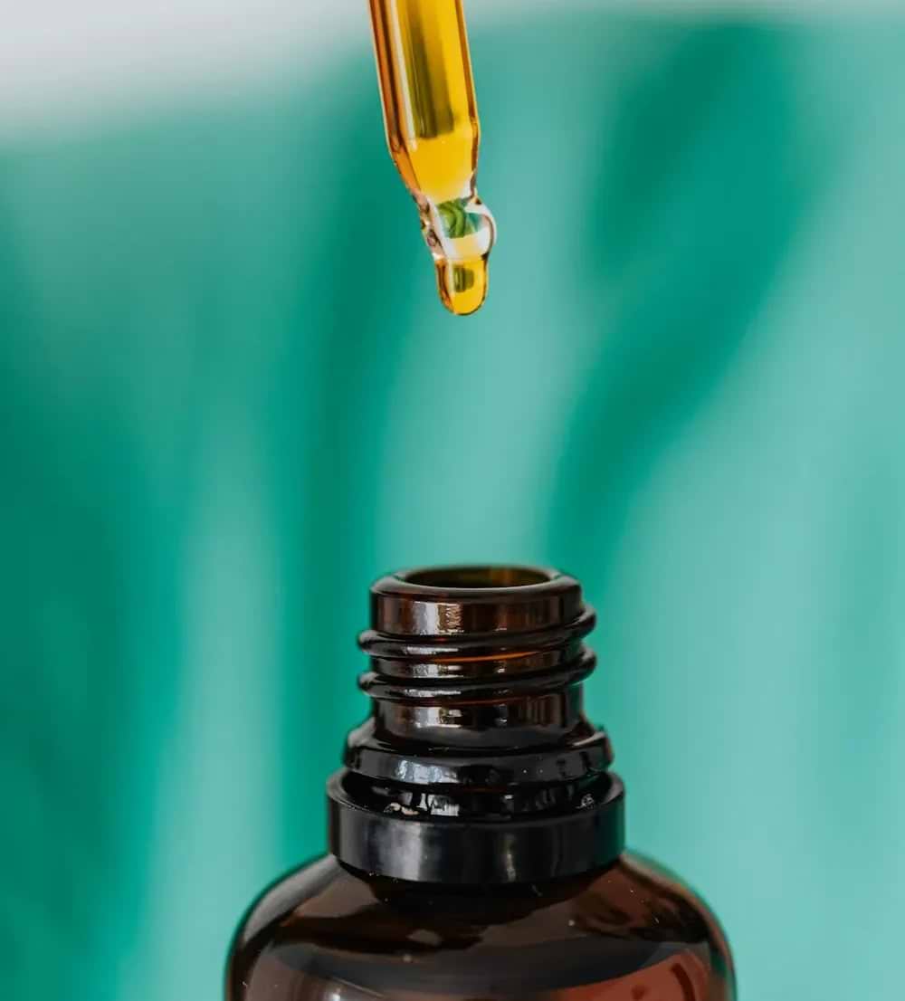 A close-up of a golden dropper releasing liquid into a brown glass bottle, set against a soft turquoise background.
