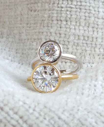 Two bezel set diamond rings, one with a yellow gold band and the other with a white gold band, positioned together on a textured fabric.