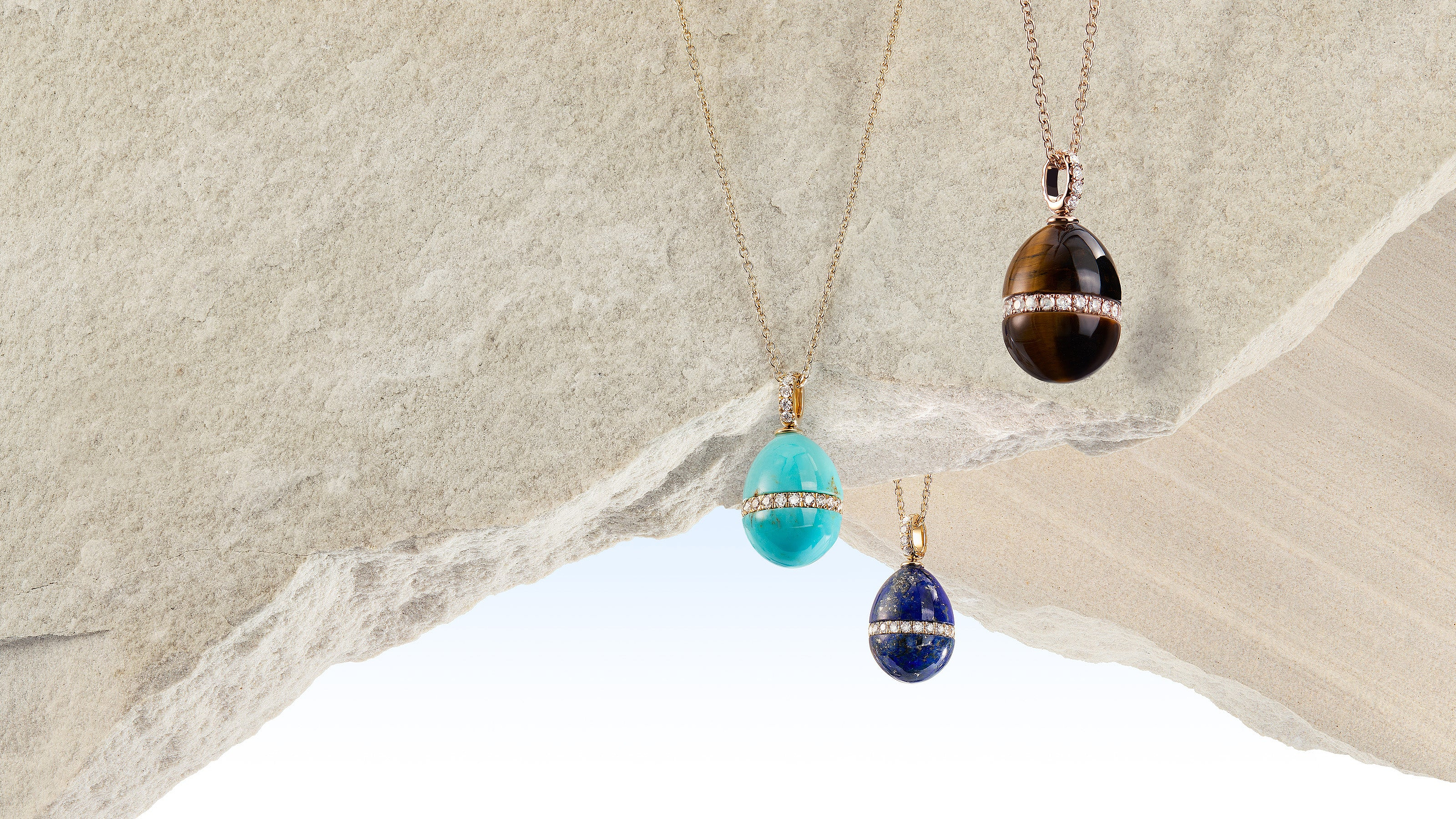 Three Fabergé necklaces with different coloured hardstone pendants hanging against a stone wall.