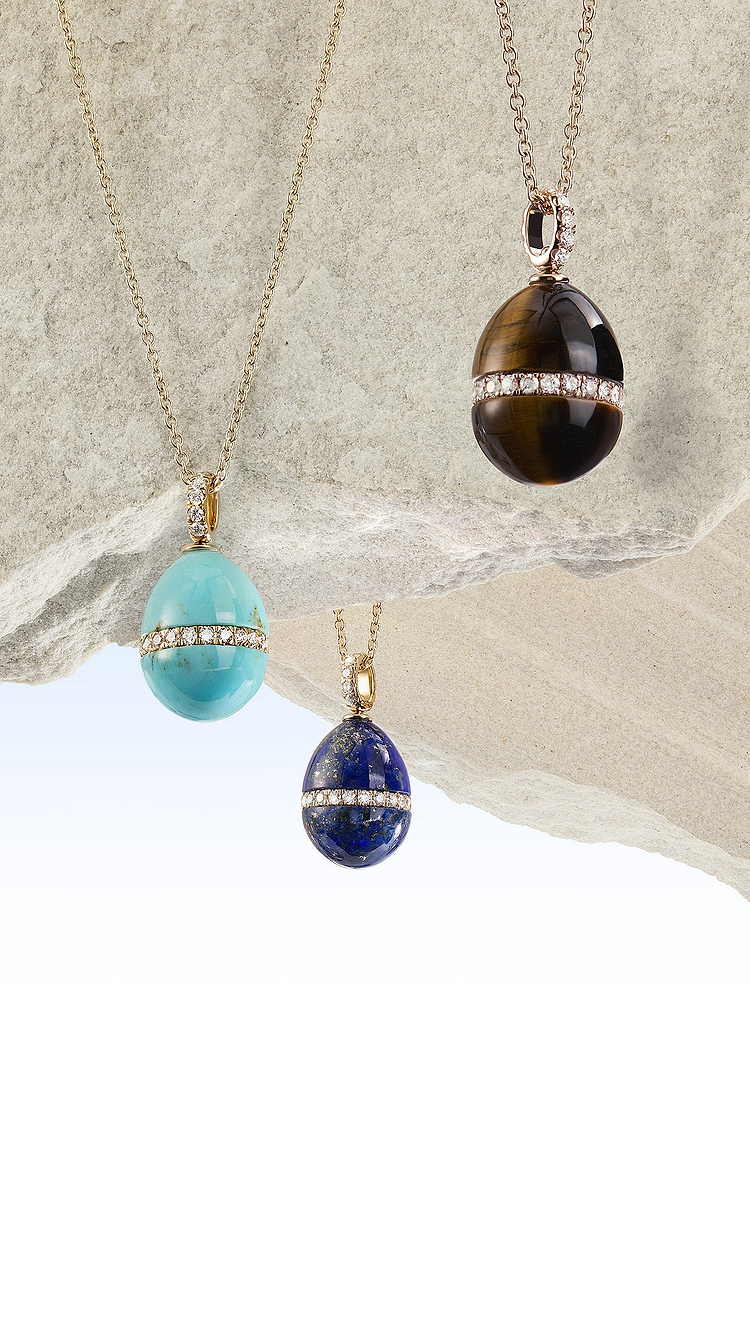 Three Fabergé necklaces with different coloured hardstone pendants hanging against a stone wall.