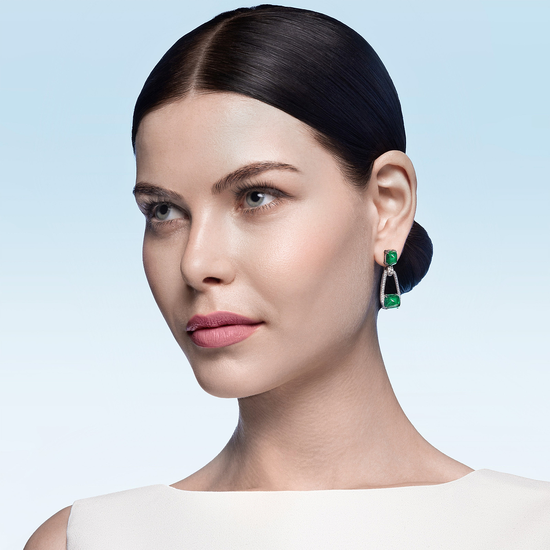 High Jewellery category - Woman wearing green emerald and white gold earrings against a light blue background