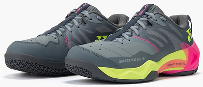 A pair of Yonex Subaxia GT WIDE Unisex Badminton Shoes in grey with neon yellow soles, pink accents, black laces, GRPHT THRTTL support, "SUBAM A1" text, Yonex logo, and a textured grip on the toe and sole.