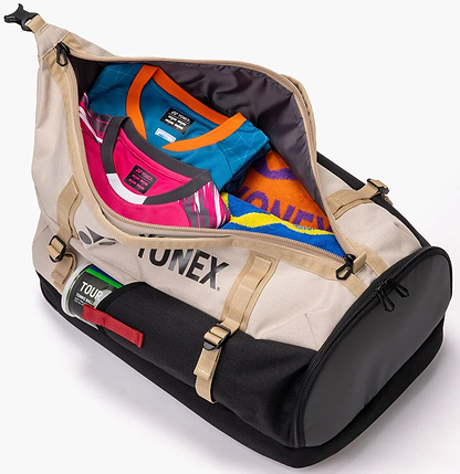The Yonex BA72612EX Gearlogic Badminton Backpack in beige is shown unzipped, revealing colorful shirts and an orange-blue towel inside. This 40L Yonex bag features sturdy straps, buckles, and branded tags on the exterior.