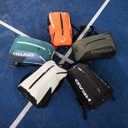 Five HEAD Tour Badminton Backpacks, each featuring a roomy shoe compartment, are arranged in a circle on a blue court with white lines. The backpacks come in orange, teal, black, olive green, and white and prominently display the "HEAD" logo.