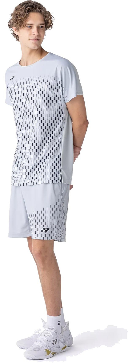 A young man stands sideways, wearing a light gray athletic t-shirt and Yonex 15241EX Knit Men's Badminton Shorts in Silver Grey, along with white sneakers. He looks left with his hands behind his back.