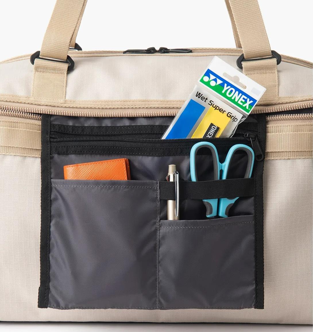 A close-up of a beige Yonex BA72611EX Gearlogic Badminton Boston Bag’s open front pocket, made from recycled polyester, neatly organizing a Yonex wet super grip pack, blue scissors, pen, and orange wallet in separate compartments.