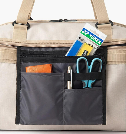 A close-up of a beige Yonex BA72611EX Gearlogic Badminton Boston Bag’s open front pocket, made from recycled polyester, neatly organizing a Yonex wet super grip pack, blue scissors, pen, and orange wallet in separate compartments.