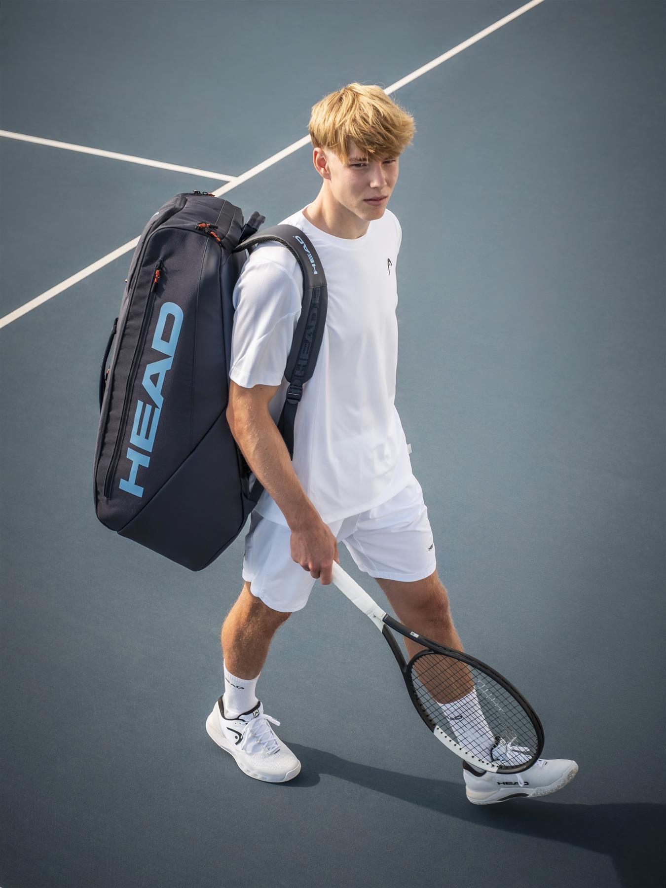 A young man in white tennis attire stands on a blue court, carrying the HEAD Pro 12 Racket Badminton Bag XL - Navy with CCT+ climate control technology on his shoulder and holding a tennis racket in his right hand.