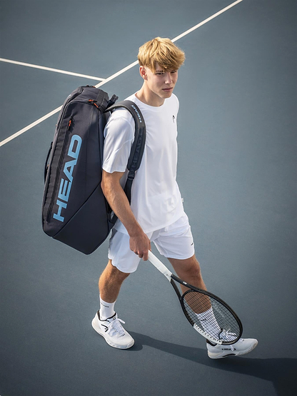 A young man in white tennis attire stands on a blue court, carrying the HEAD Pro 12 Racket Badminton Bag XL - Navy with CCT+ climate control technology on his shoulder and holding a tennis racket in his right hand.