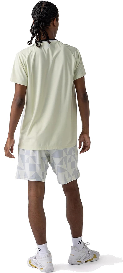 A person with medium-length braided hair stands facing away, wearing a light green short-sleeve shirt and Yonex 15240YX Knit Men's Badminton Shorts in Champagne, along with white socks and white athletic shoes on a white background.