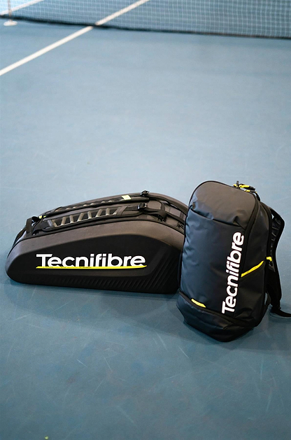 Two black Tecnifibre Reform L Badminton Racket Bags with white Tecnifibre logos and yellow accents rest on a blue tennis court near the net. These bags are spacious and made with durable EVA material.