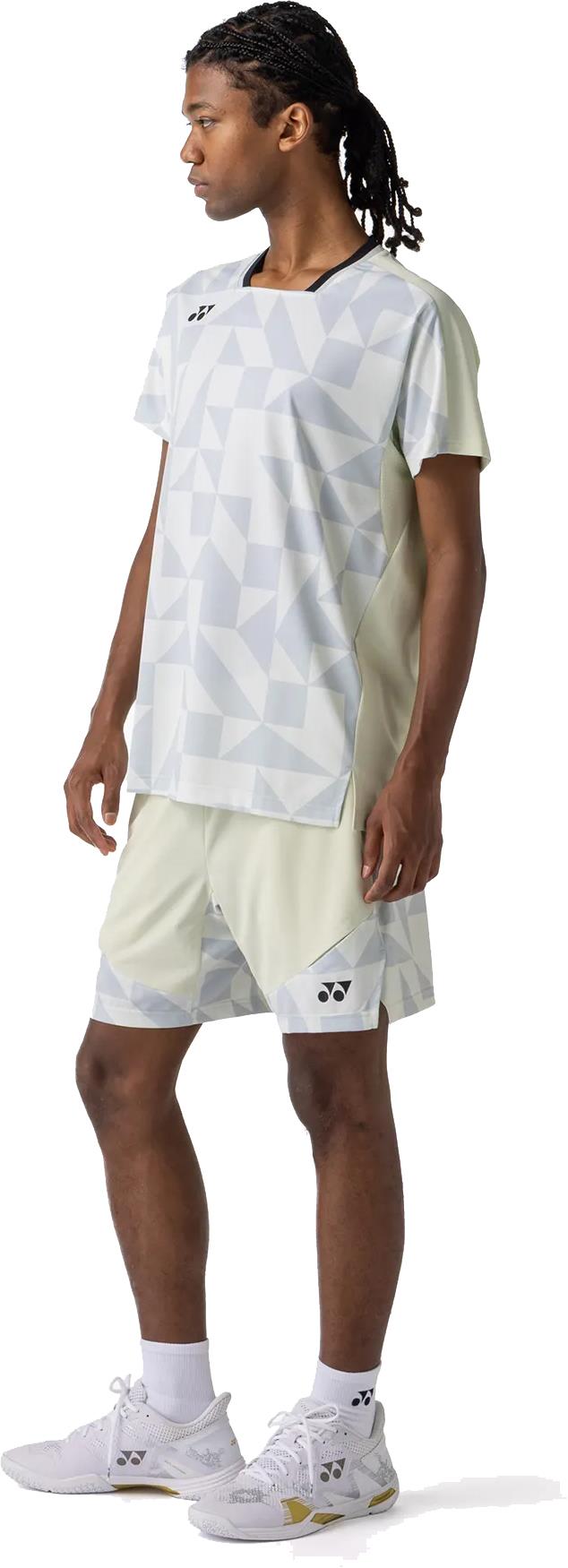 A person stands in profile on a white background wearing a light geometric athletic shirt, white socks, sports shoes, and Yonex 15240YX Knit Men's Badminton Shorts in Champagne by Yonex.