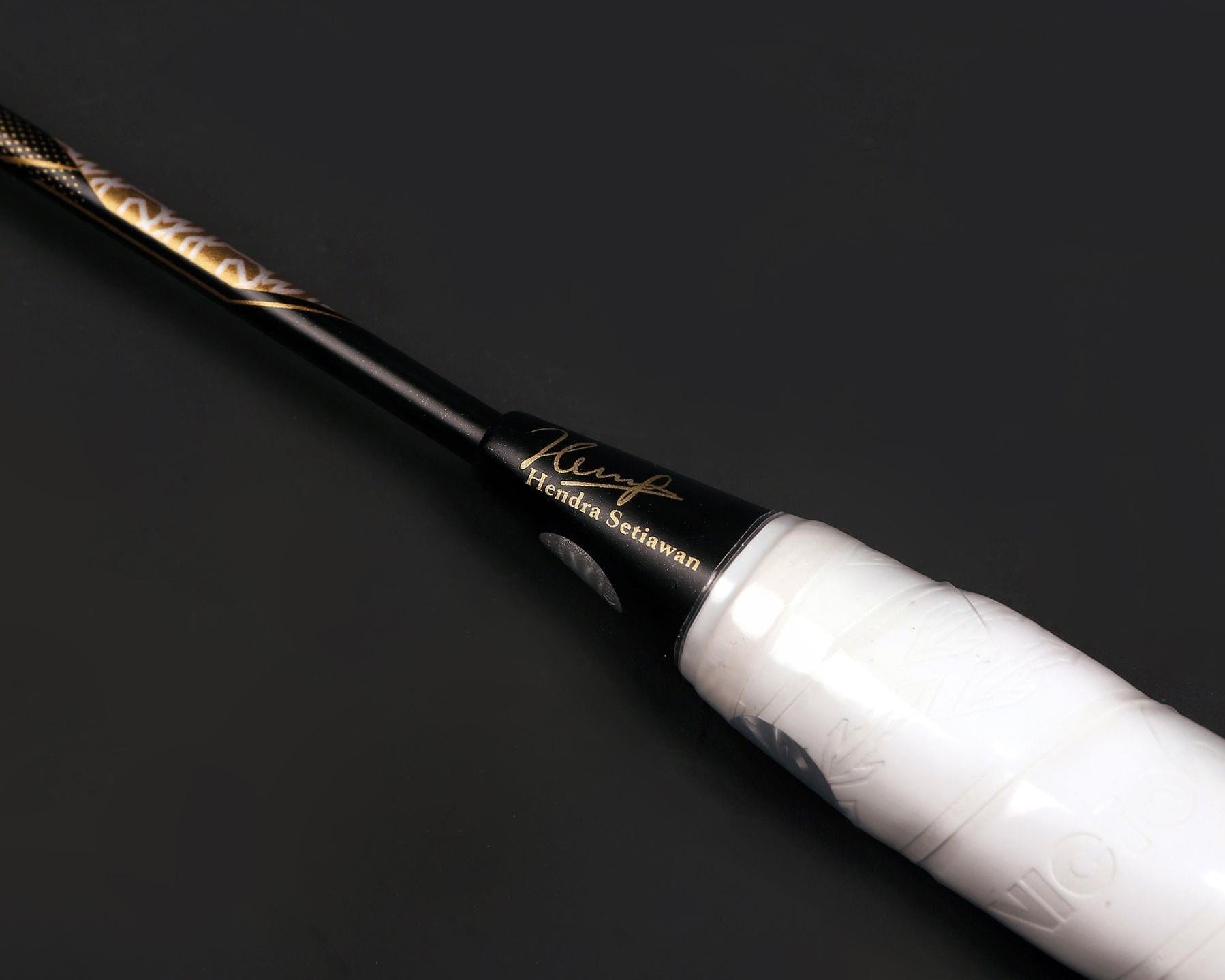 Detailed view of the Victor Thruster FC Hendra Setiawan LTD Edition 3U Badminton Racket handle, with a white grip highlighting Hendra Setiawan's signature in gold against a black background as part of the Thruster F series.