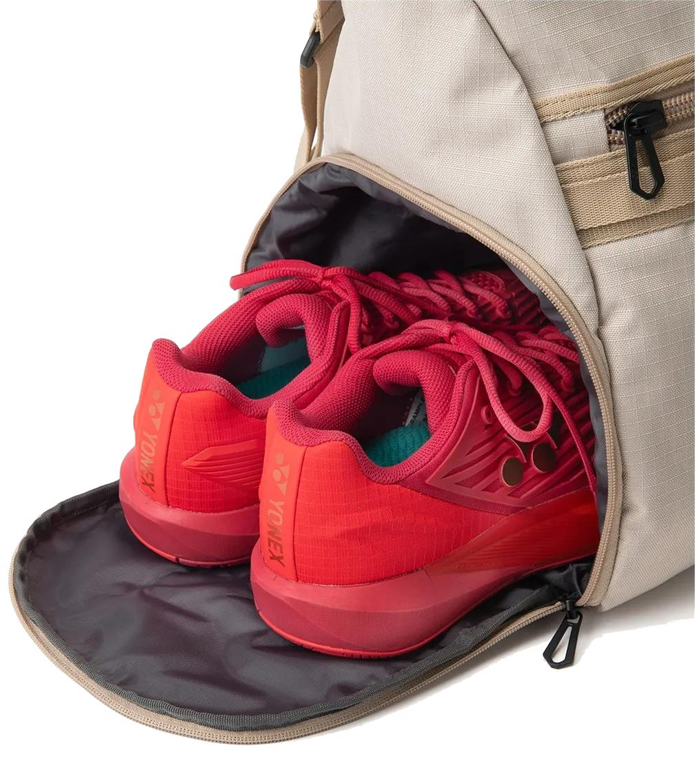 A pair of bright red Yonex shoes is stored in the zippered shoe compartment of a partially open Yonex BA72611EX Gearlogic Badminton Boston Bag (Beige), revealing the shoes inside.