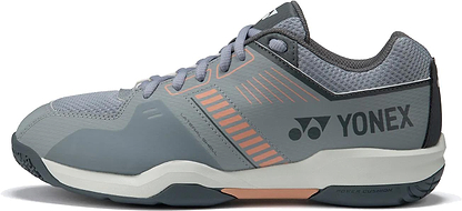 The Yonex Strider Flow Unisex Badminton Shoes in light grey feature light orange accents, breathable mesh, and a cushioned sole—ideal for beginners. Pictured from the side against a white background.