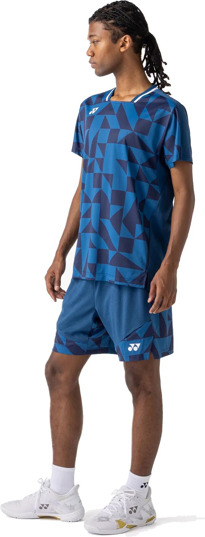A person stands sideways in a blue geometric-pattern sports shirt and Yonex 15240YX Knit Men's Badminton Shorts - Indigo Blue, with white socks and athletic shoes, looking left against a plain white background.