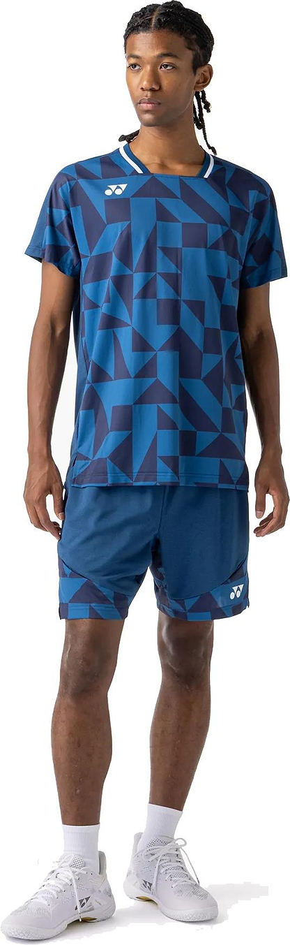 A person wears the Yonex 10712YX Crew Neck Men's Badminton T-Shirt in indigo blue with geometric patterns, paired with matching shorts, white socks, and white athletic shoes against a plain white background.