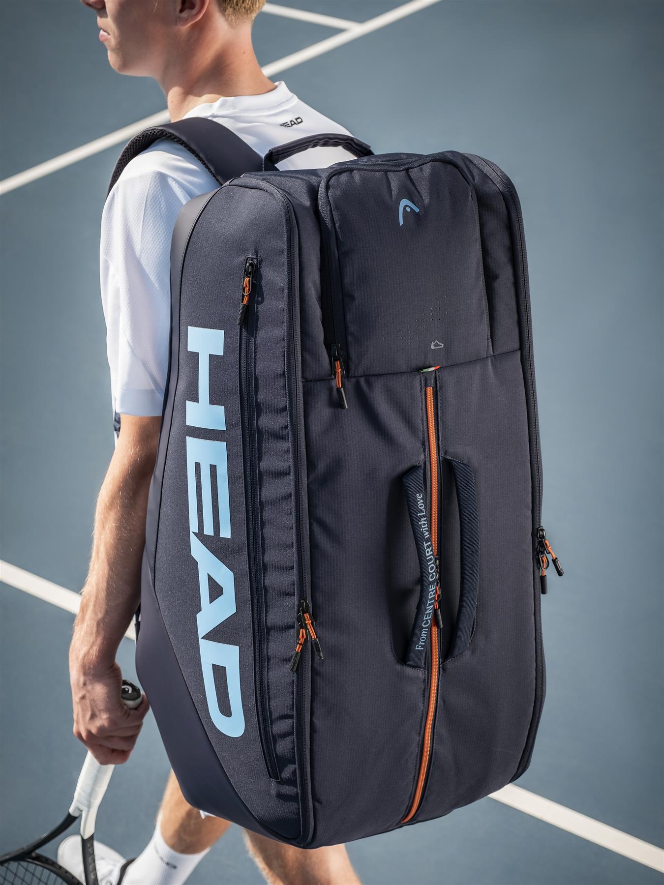 A tennis player stands on the court with a HEAD Pro 12 Racket Badminton Bag XL in navy on their back, featuring CCT+ climate control technology, and holds a racquet in one hand.