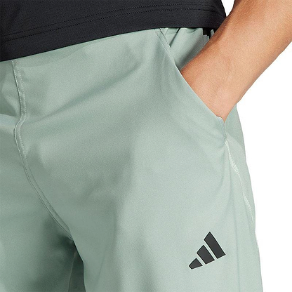 A person is wearing green ADIDAS Melbourne Pro Badminton Pants with a black logo on the left leg, standing with their hand in the pocket. Only part of their torso and arm are visible, paired with a dark shirt.