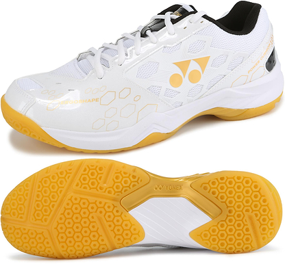 The Yonex Power Cushion SHB 101 Badminton Shoes in white and gold feature honeycomb patterns, a yellow textured grip sole with shock absorption, and the Yonex logo on both the side and sole.