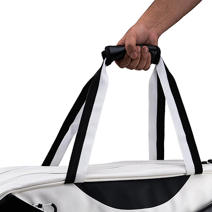 A person's hand holds the Li-Ning Competition 6 Racket Badminton Bag in white with black accents, a duffel-style bag by Li-Ning designed to carry up to 6 rackets, shown against a plain white background.