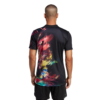 A man with short hair is wearing an adidas Melbourne Men's Freelift Polo Shirt in black and multi colors, which features a colorful abstract pattern on the back with vivid shades of red, yellow, and blue. He pairs it with black shorts and stands against a plain white background.