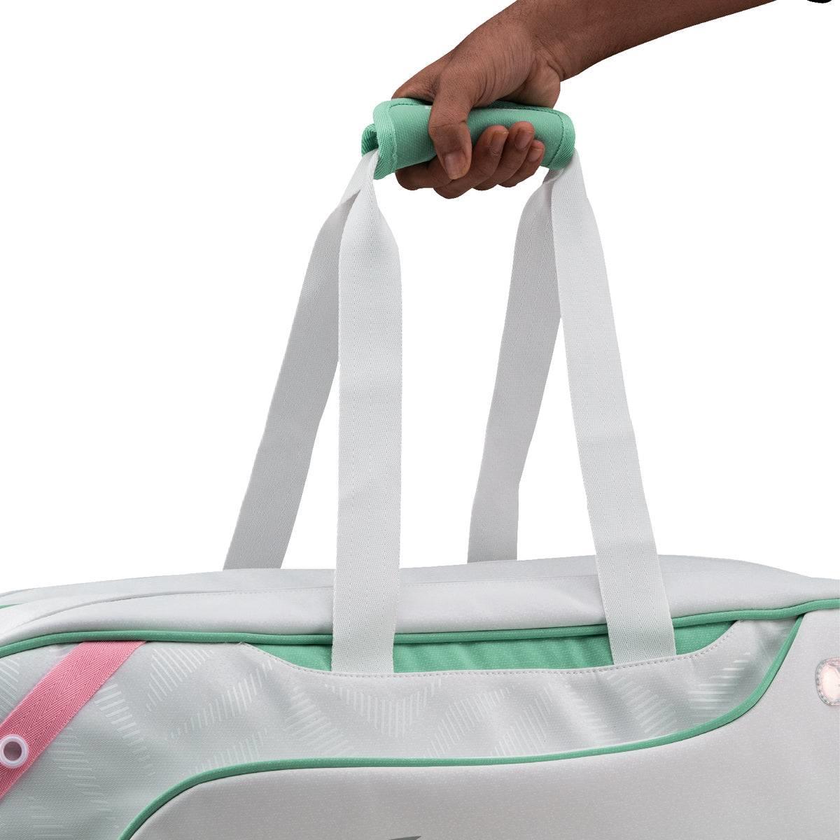 A hand holds the mint green handle of the Li-Ning Competition 6 Racket Badminton Bag in white and green, showing off its stylish upper section with white straps and a pink accent.