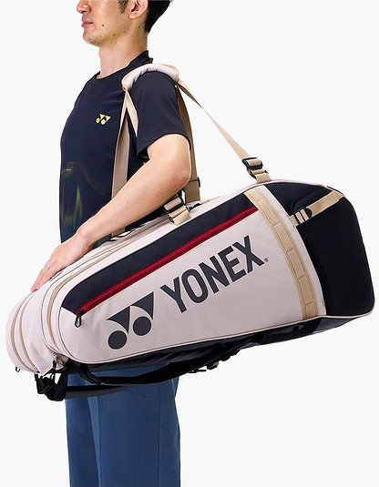 A person in a navy Yonex t-shirt carries the Yonex BA72629EX Gearlogic 9 Racket Badminton Bag in beige and black with red zipper and shoulder strap; the Yonex logo is clearly displayed on the side.