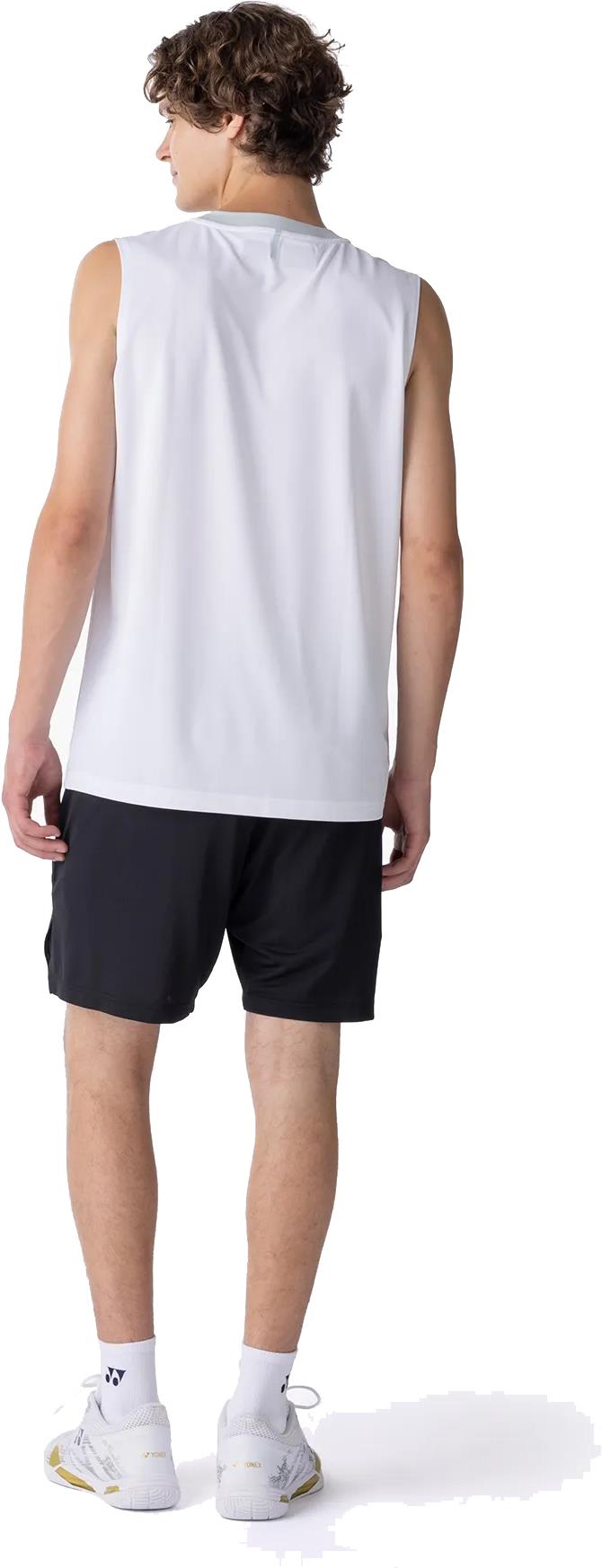 A young man with curly hair stands facing away, wearing a sleeveless white shirt, Yonex 15239YX Knit Men's Badminton Shorts in black, white socks, and white athletic shoes.