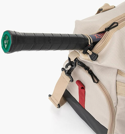 A close-up of the beige Yonex BA72611EX Gearlogic Badminton Boston Bag, made from recycled polyester, with a racquet handle in a side zip pocket. The bag has tan straps, black base, and a red accent loop.