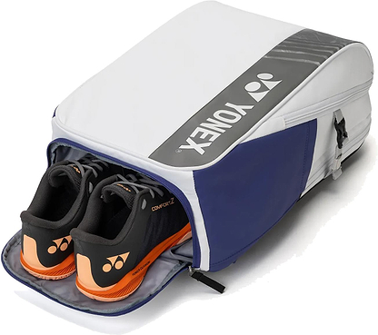 A Yonex BA52512EX Club Badminton Backpack in white and blue from Yonex is shown unzipped, revealing black and orange Yonex athletic shoes stored in a separate compartment.