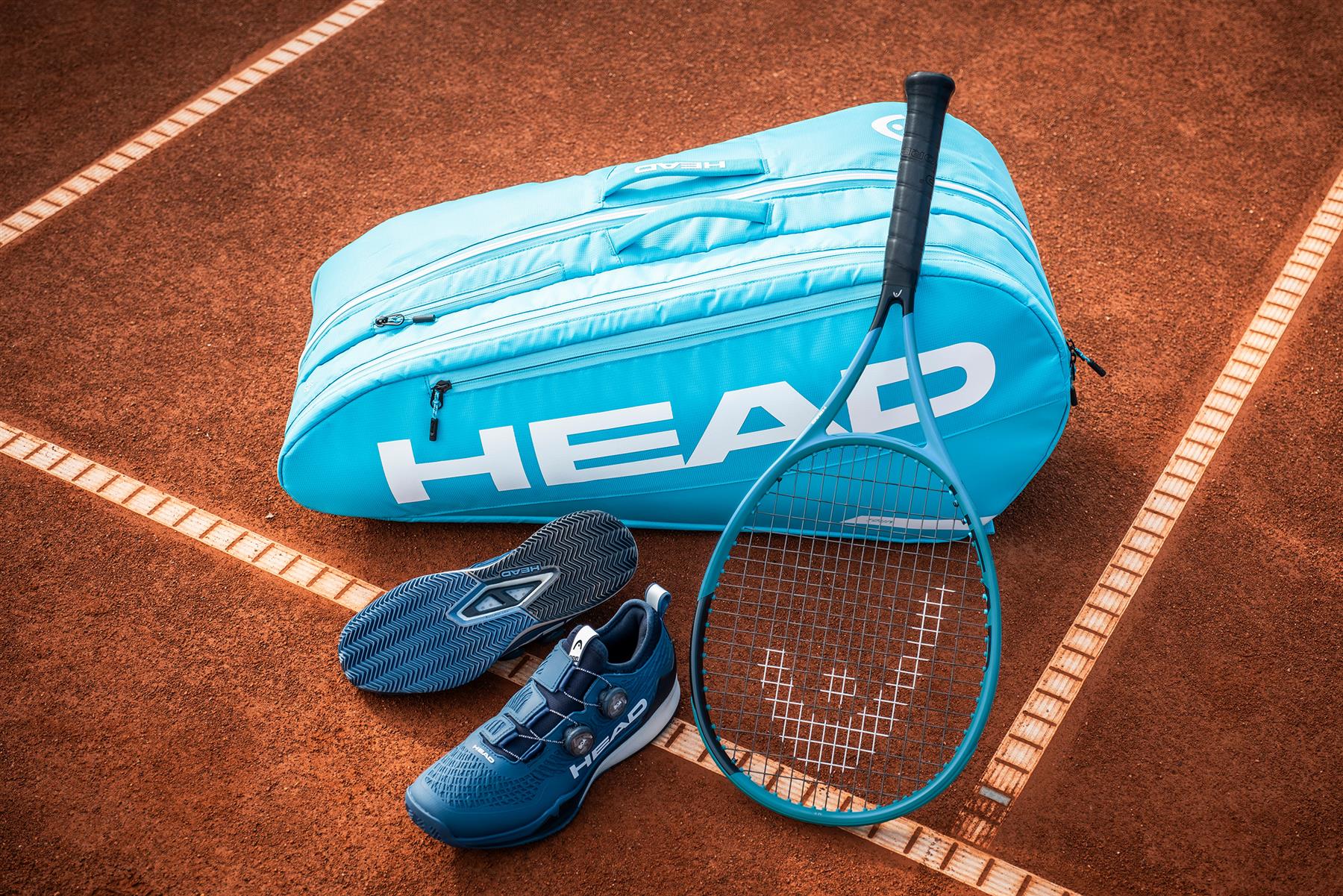 A HEAD Tour 9 Racket Badminton Bag L - Blue, a matching racket, and blue tennis shoes are placed on a clay court near the sideline.