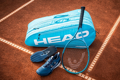 A HEAD Tour 9 Racket Badminton Bag L - Blue, a matching racket, and blue tennis shoes are placed on a clay court near the sideline.
