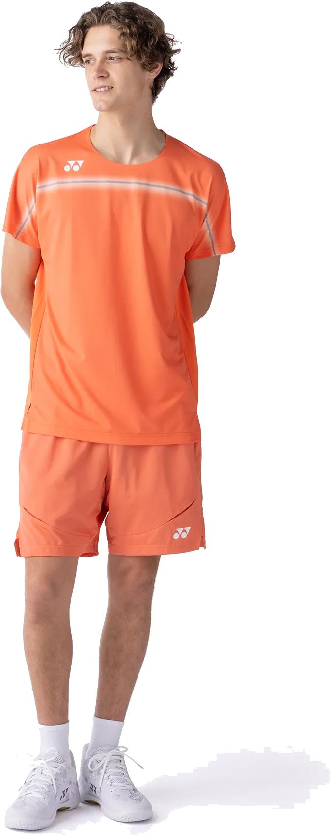 A young man with curly hair wears the Yonex 10711YX Crew Neck Men's Badminton T-Shirt in Cyber Orange, paired with matching shorts, white socks, and sneakers. He stands hands behind his back, looking to the side against a white background.